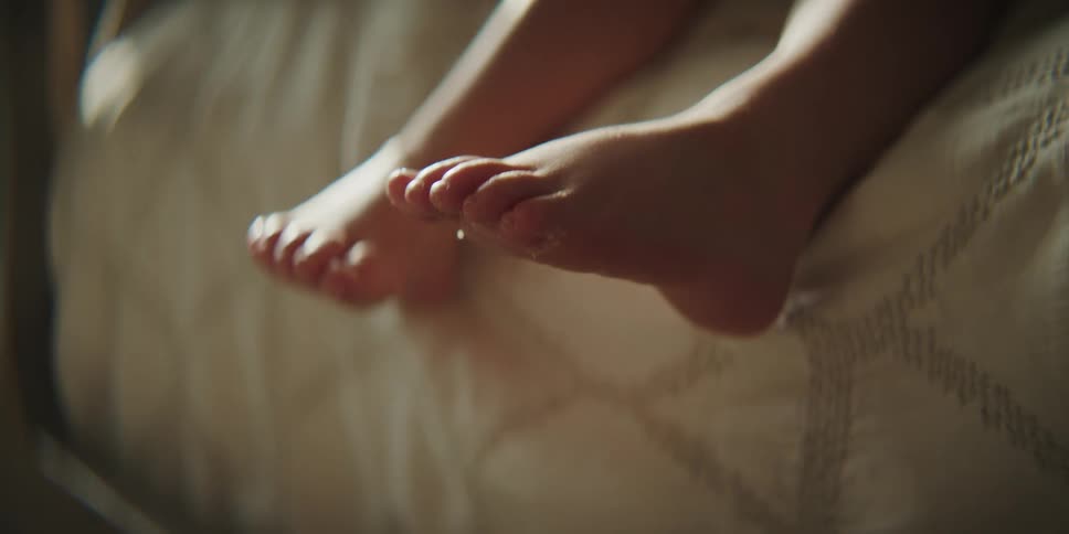 The feet of a child sitting with their legs dangling off the side of a bed