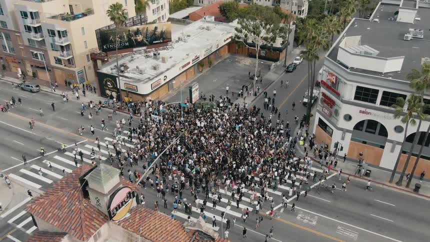 A large crowd of people stand outside stores in a city during a protest