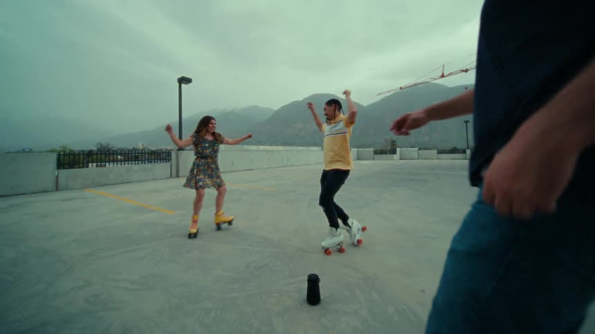 Two men and a woman dance on roller skates on a roof top building with a construction crane and hills in the background
