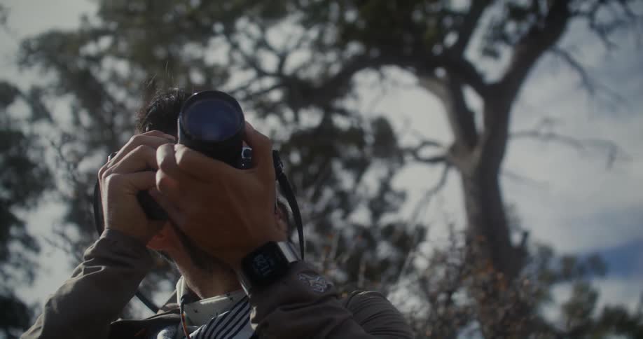 A man is standing outside near trees and holding a camera to his face as he turns the lens