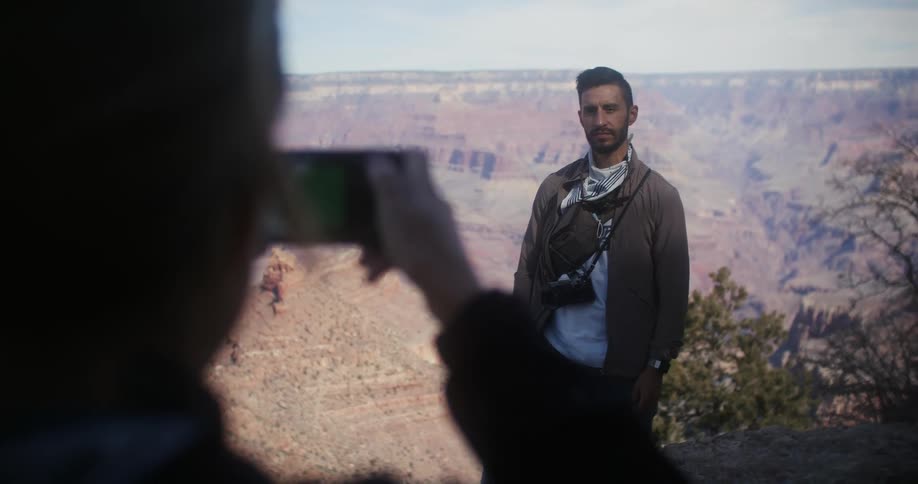 A woman uses a smartphone to take a photograph of a man posing on the edge of an immense canyon during the day