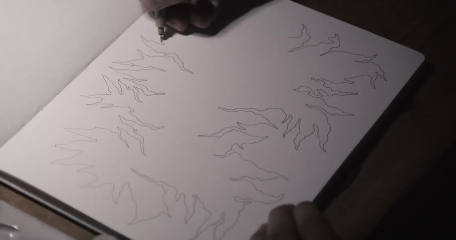 A man is sitting down at a table drawing on paper with a black pen