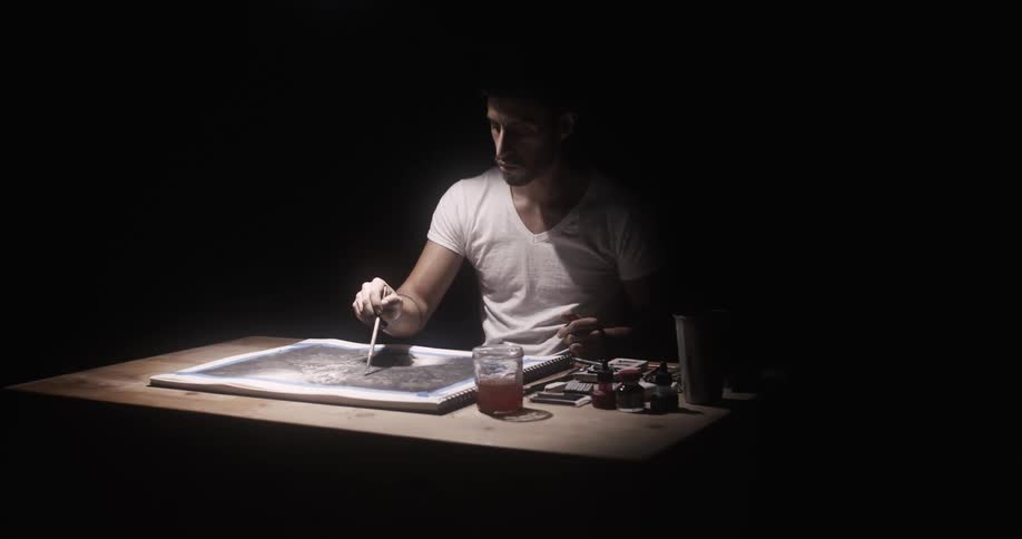 A man is sitting in the dark at a desk painting on a canvas