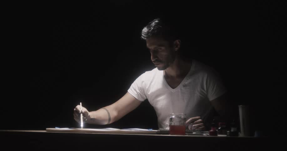 Concentrated painter sitting at the desk in a dark room painting a sketchbook with a painting brush