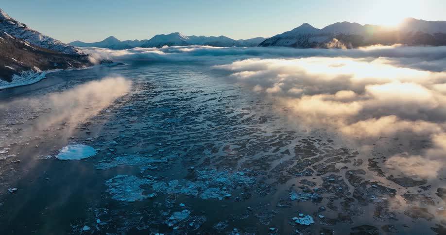 Fog lays low over a partially frozen body of water in the snowy mountains