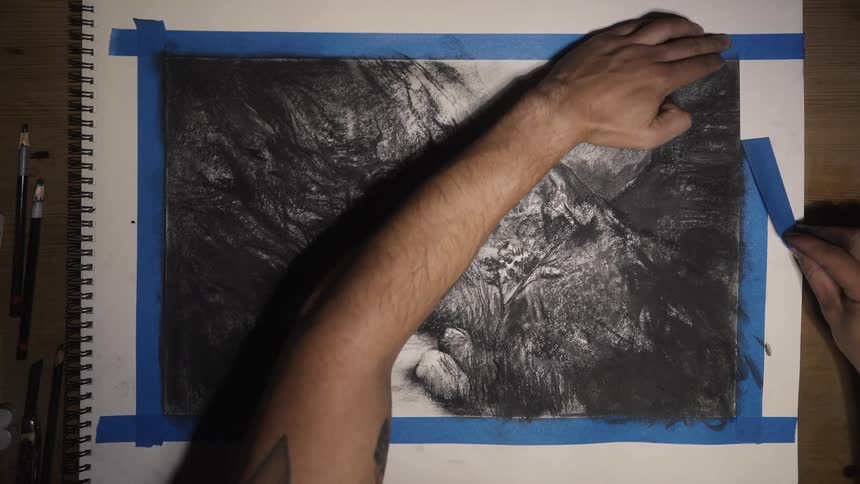 Artist slowly pulls off blue tape leaving clean edge on inked drawing in sketchpad