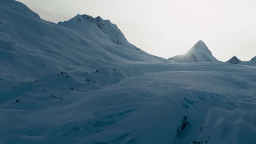 The sun shines behind snow covered mountains with peaks high in the sky