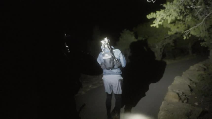 A pair of men use flashlights to light their way while hiking along a pathway at night