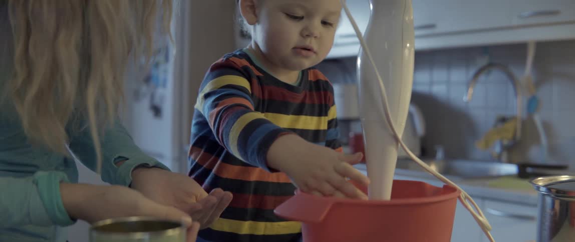 A mother and young toddler prepare food together in a bowl at a kitchen counter.