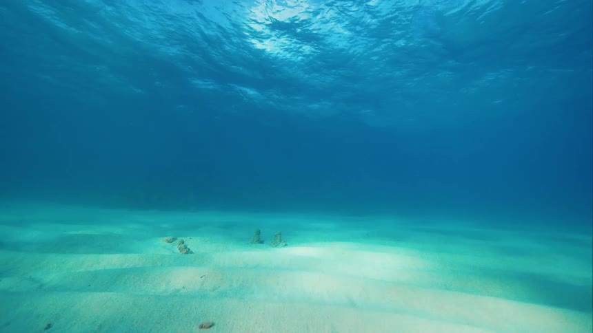 Underwater view of the sun shining through clear blue water and ripples reflecting on the seabed below