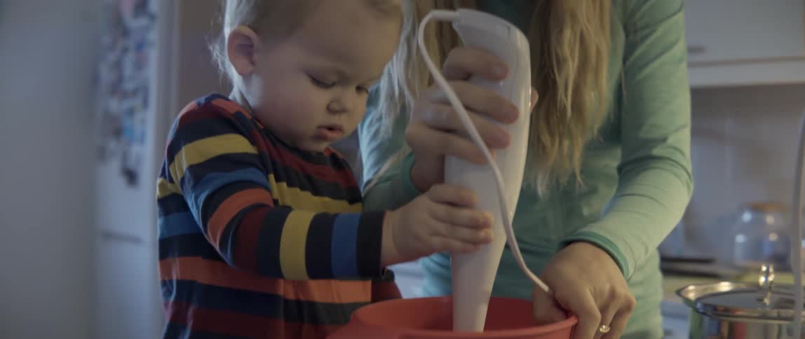 A toddler watches and helps a woman using a hand mixer
