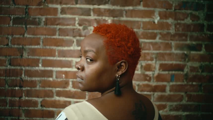 A woman faces a brick wall turns to look into the camera