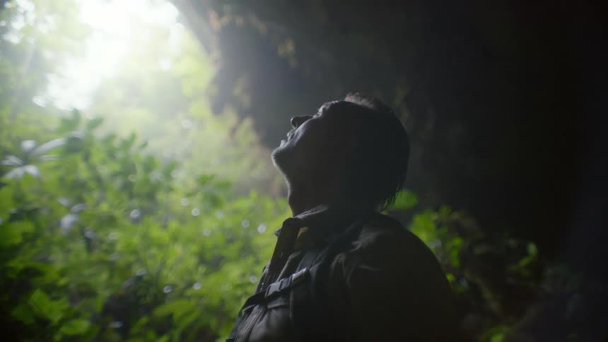 A man out hiking stands under a cliff in a forest and looks up