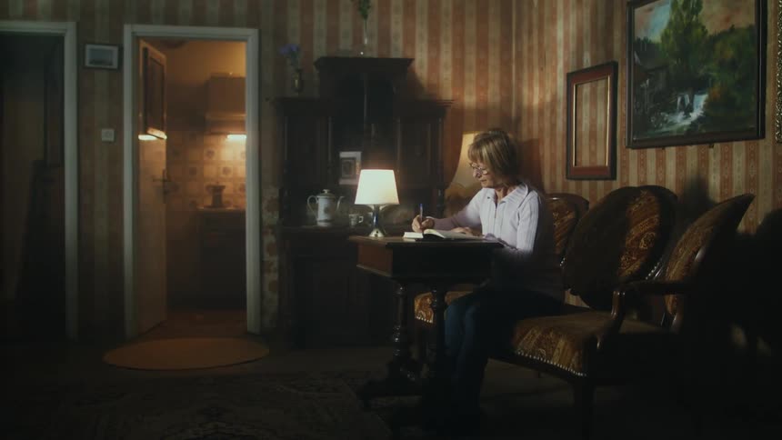 An elderly woman is sitting at a table writing in a book and there is an empty picture frame on the wall behind her
