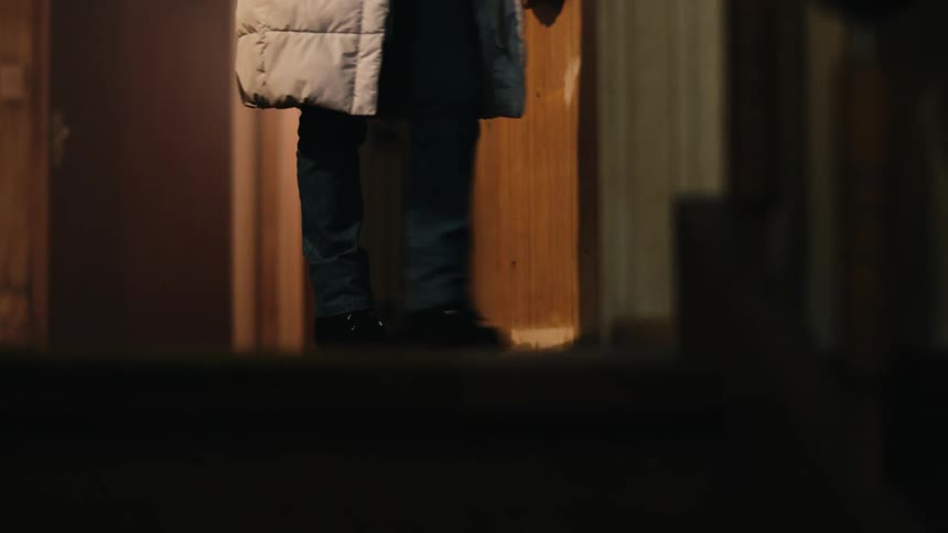 A woman wearing a coat walks down a house hallway and into another room