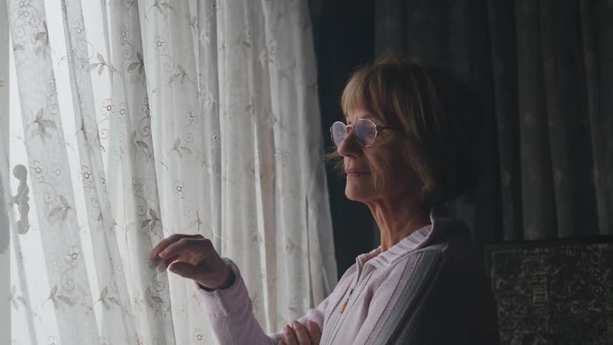 An elderly woman at home peels back the curtains and looks outside