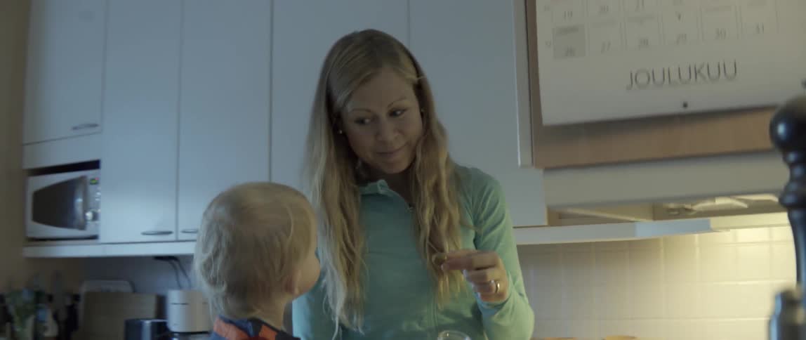 A mother feeds a young toddler an olive while preparing food in the kitchen.