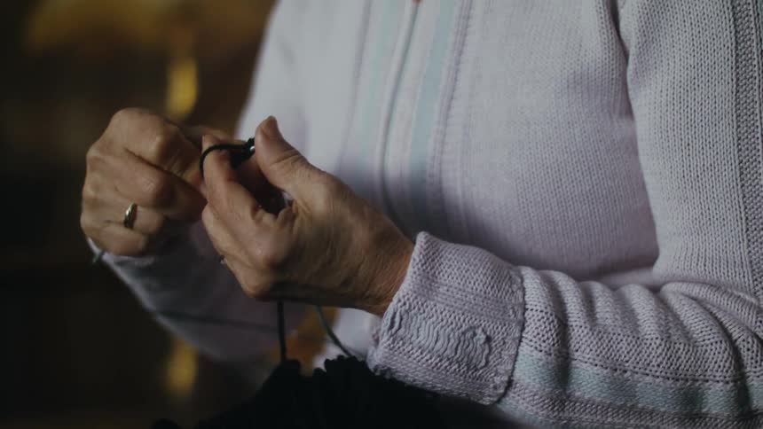 A woman uses a large needle to knit while sitting indoors