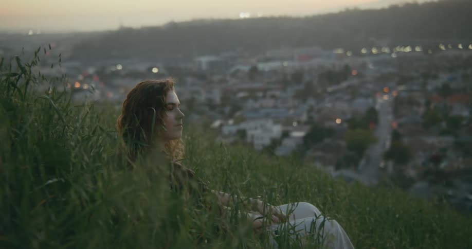 A woman looking out over a city from a hillside on the outskirts lays back into the grass