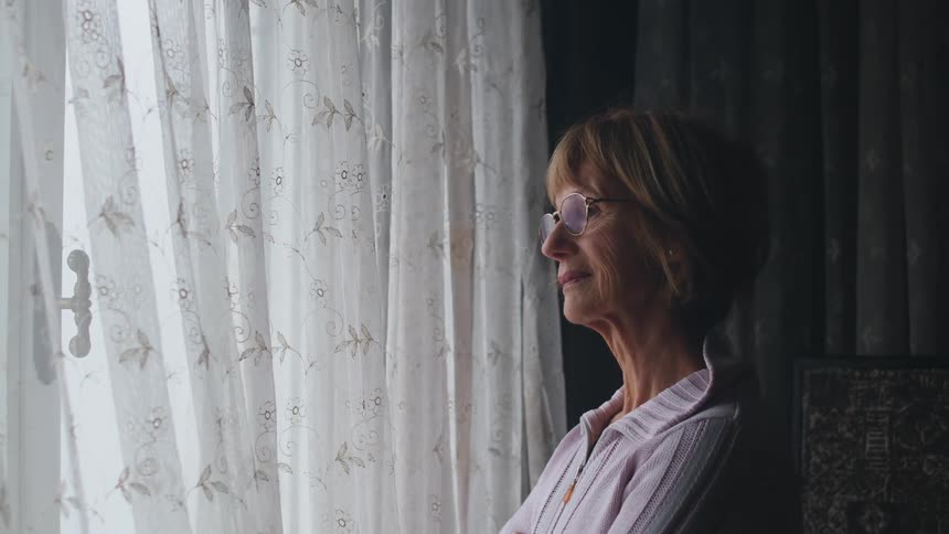 An elderly woman standing next to a curtained window