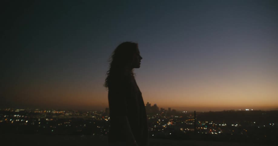 A teenage girl stands on a hill and stares in to the distance with a city skyline below her in the distance