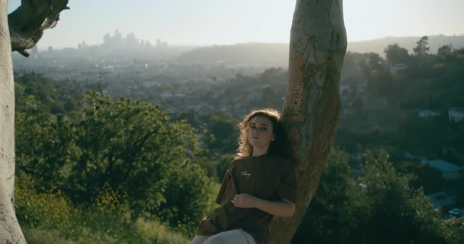 A woman leaning back against a tree on a slope overlooking homes in a valley and a city skyline on the horizon