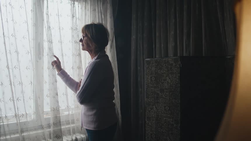 An elderly woman is standing at her window and she lifts the curtain to look outside and the lamp is on in the daytime