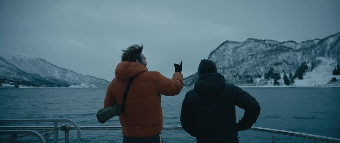 A pair of people gesture at the snowy coastline in the distance while standing on the deck of a boat on a cloudy day