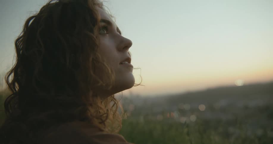 A young woman looks up at the sky while sitting in the grass on a hillside outside of a city in the evening