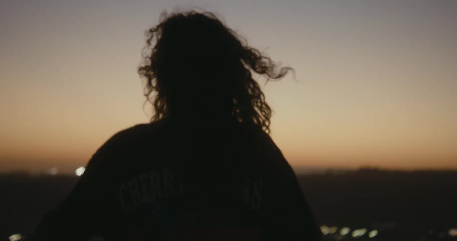 A woman twirls and moves around aimlessly while standing on a rooftop in a metropolis at sunset