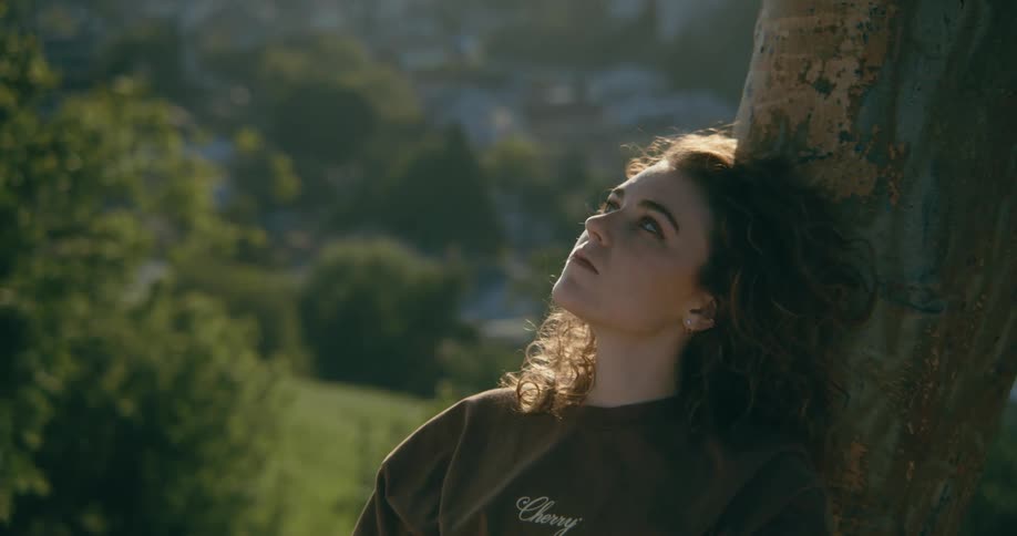 A woman gazes up at the sky while resting against a tree during the day