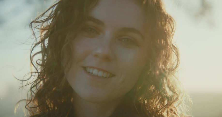 Woman with curly hair and freckles smiles at camera at sunset