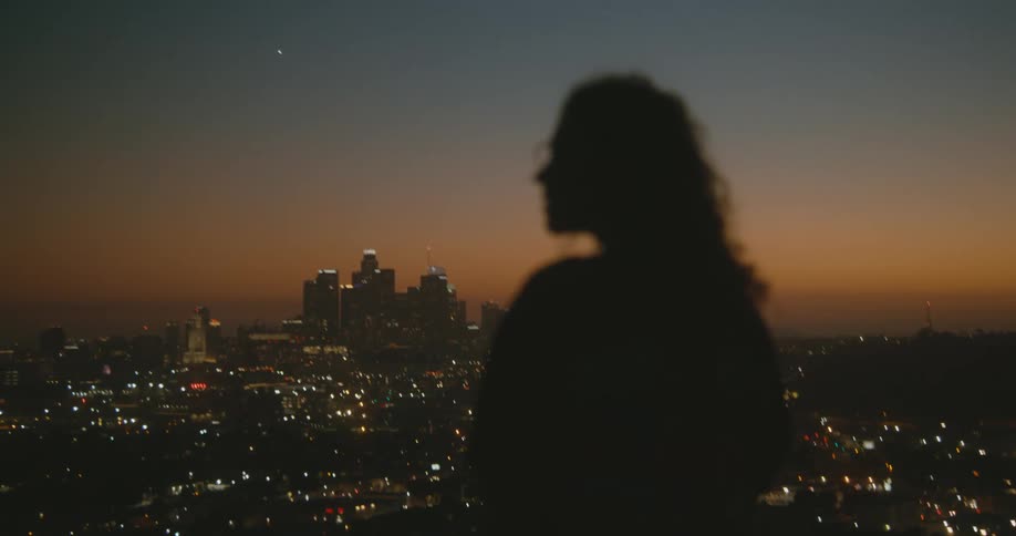 A woman runs her hand through her hair while looking out over a city from a rooftop at night