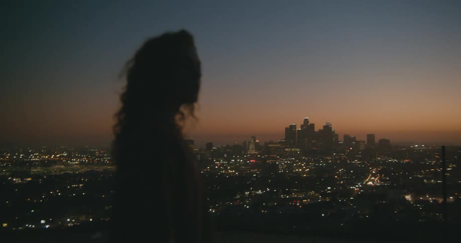 A woman is standing outdoors near a downtown city while the sun sets