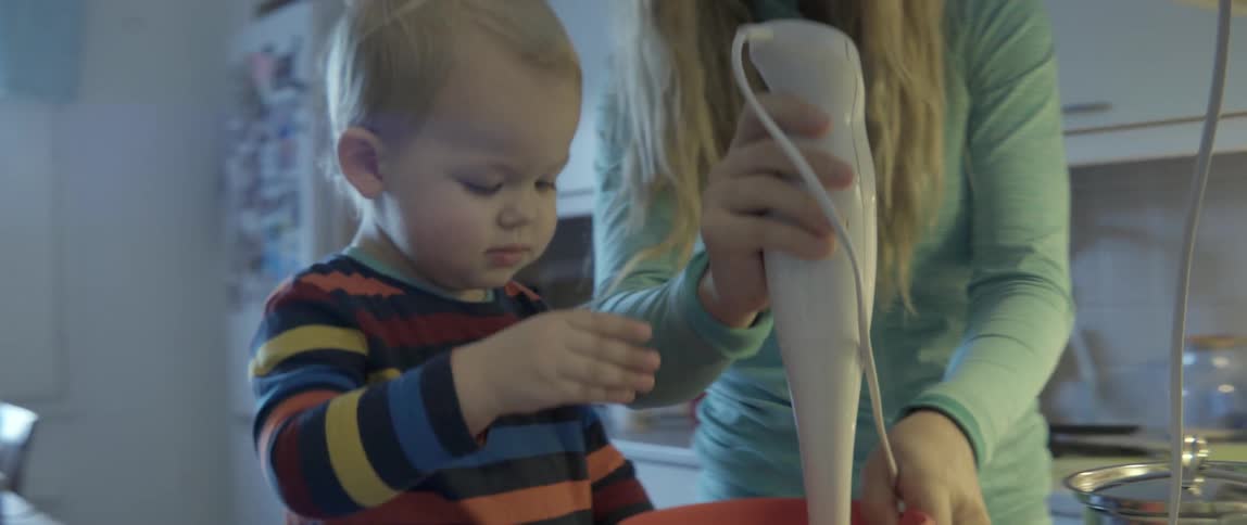 A young toddler watches as his mother uses a hand blender in a kitchen.