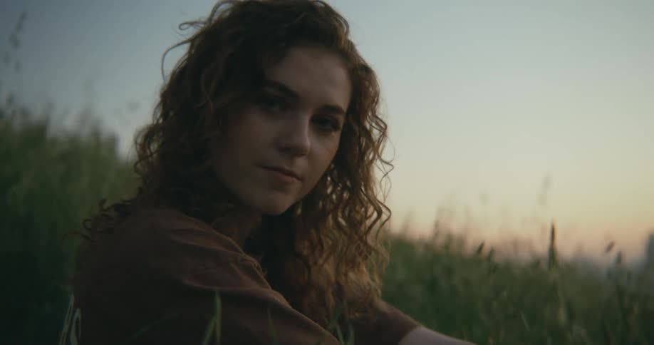 A woman looks into the camera while sitting in tall grass on a hillside at sunset