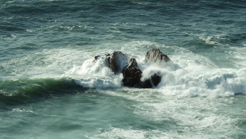 Waves roll in and crash against rocks in the water and splash high in the air