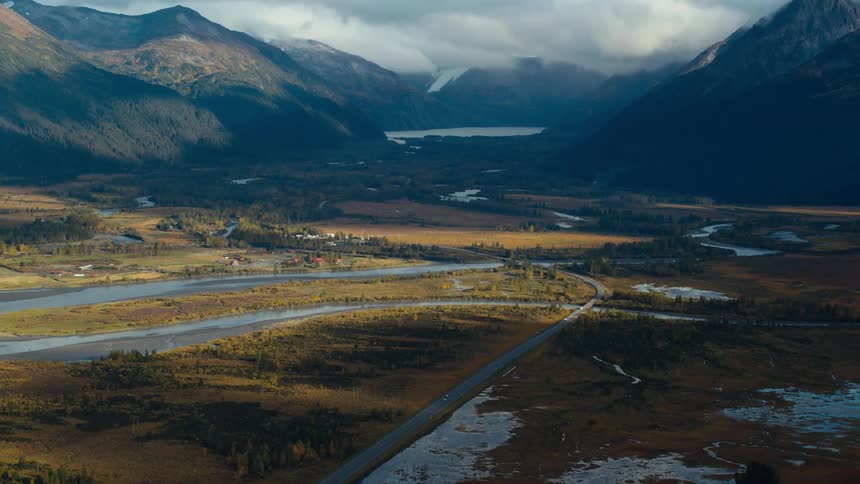 Cars move along roadthrough valley passing rivers streams and lakes with misty mountains in the background