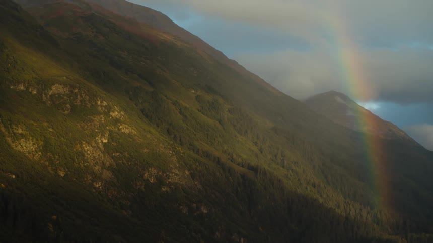 A rainbow comes down on the side of a mountain and a mountain with a peak high in the clouds is in the distance