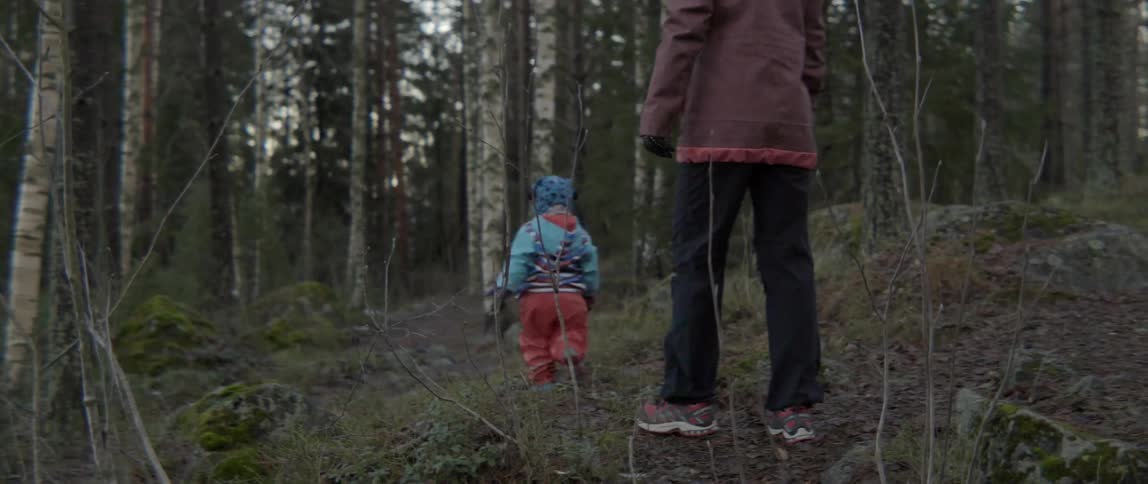 A child and woman are hiking on a hilly area towards a forest