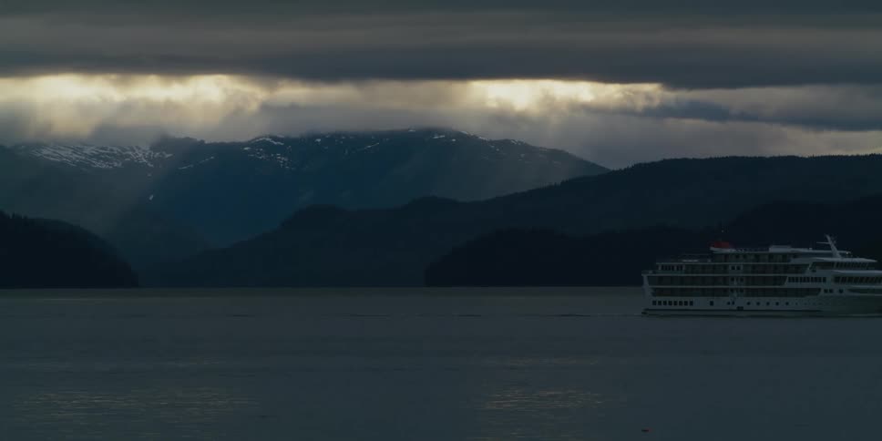 A cruise ship travels near a mountainous coastline as thick clouds darken the sky during the day