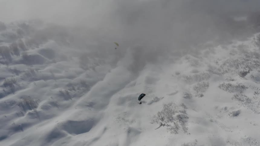 Two people paragliding down a snow covered mountain at a cloudy day