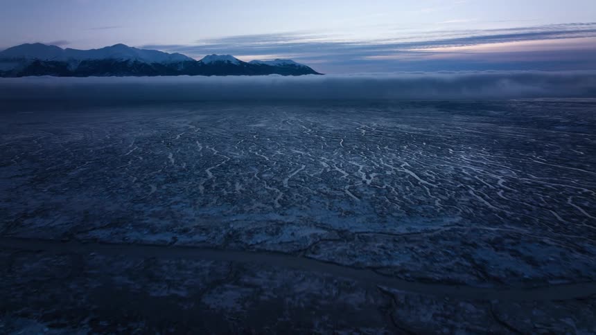 Icy tundra stretches to snow covered mountains as low lying clouds flow across the landscape under a red sky