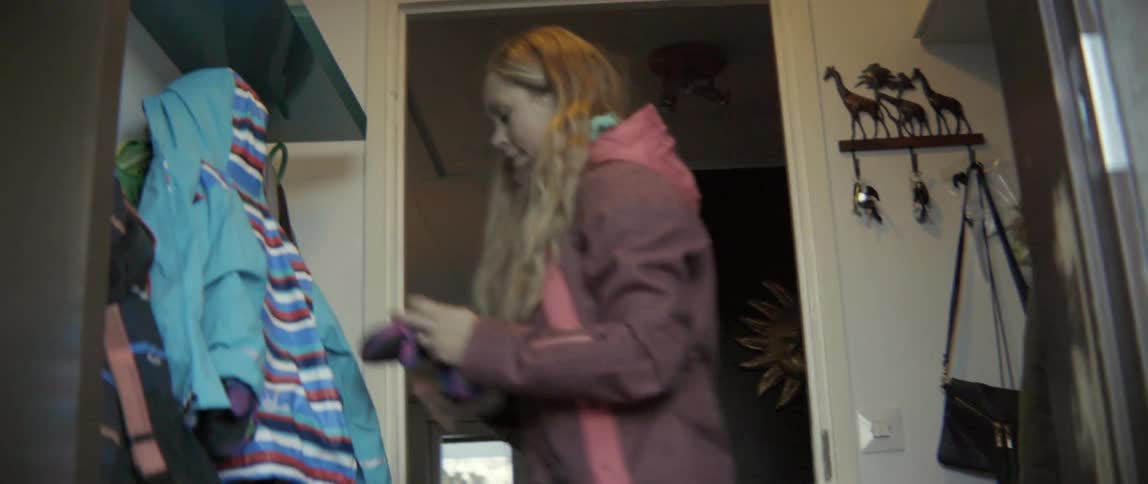 A woman collects clothes from the floor as a child walks around dropping them