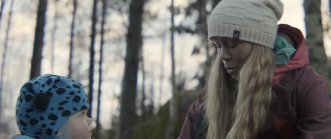 A blonde woman wearing a knit hat talks to her toddler in the woods.