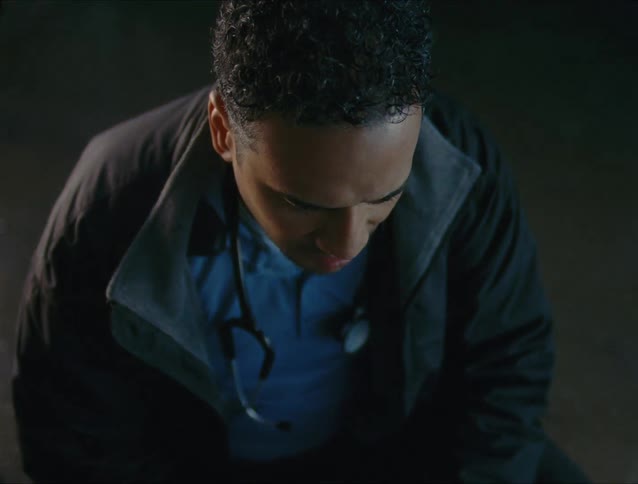 Medical responder wearing a stethoscope crouches on road under flashing red and blue lights talking to someone