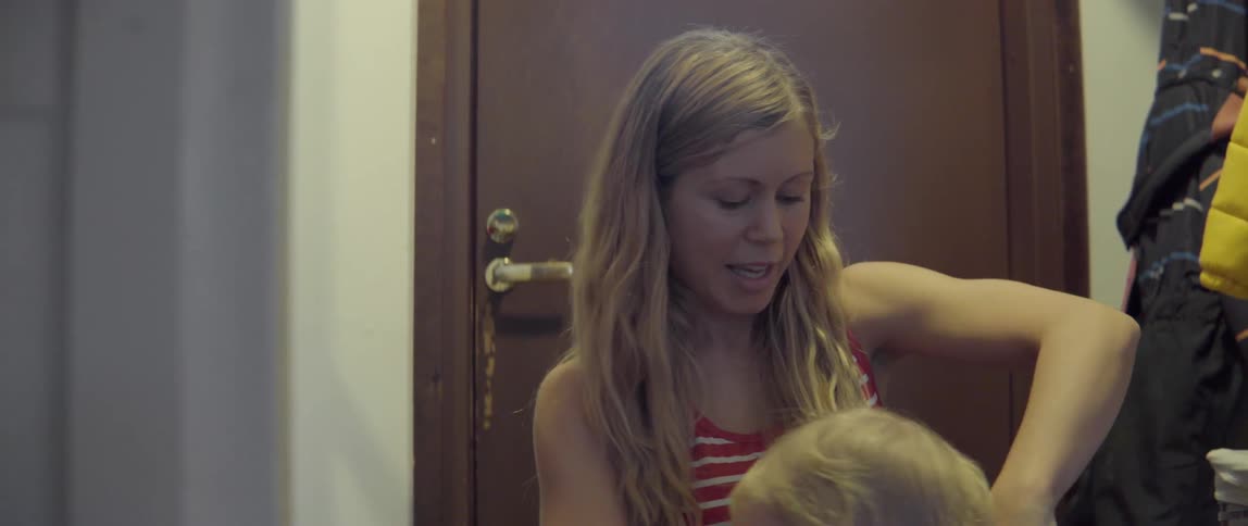 A blonde woman sits on the floor interacting with her toddler in a hallway.