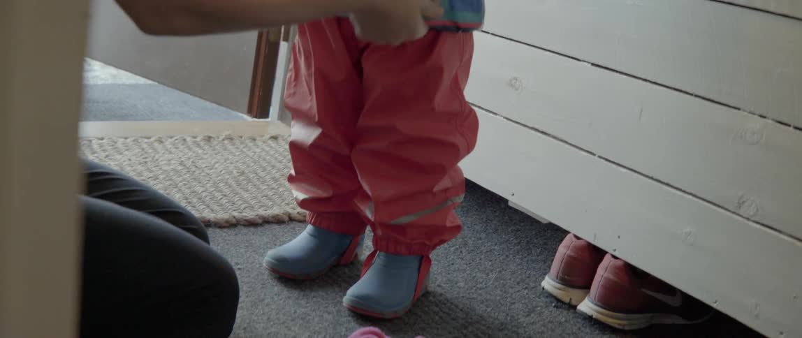 A woman assists a young toddler in putting on warm winter clothing at home.