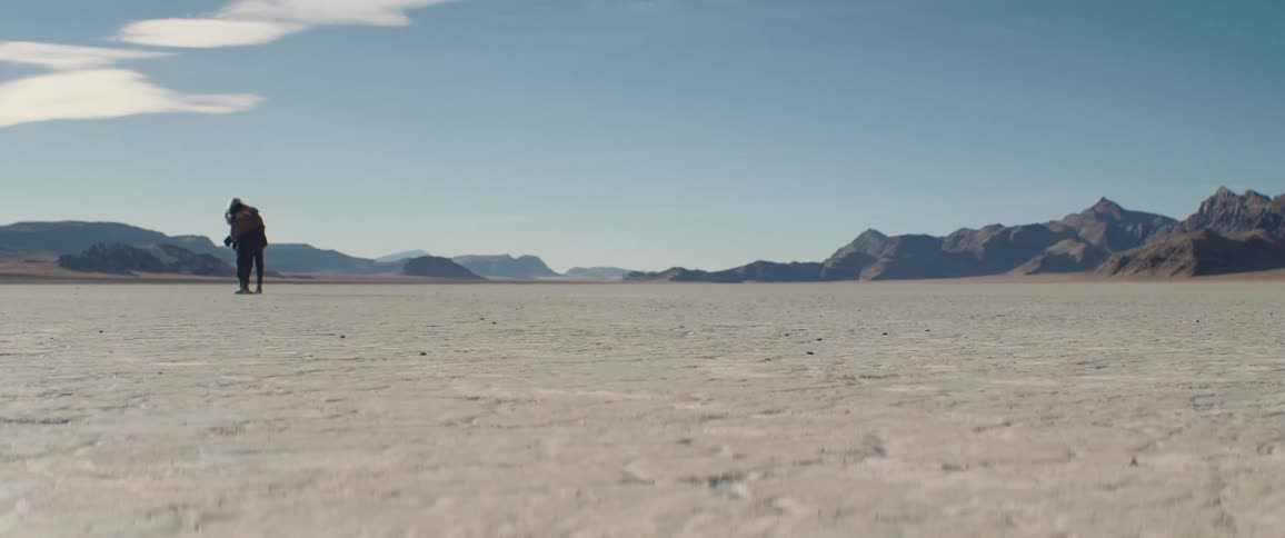A woman is standing in an open desert area surrounded by mountains with her arms open and a man runs to her and they hug