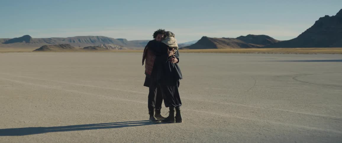 Gray haired woman comforts man with a hug as they stand in desert